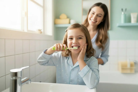 Young girl brushing her teeth with an Aussie Kids Toothbrush Co® green children's toothbrush in a bathroom, with a smiling mother standing behind her.