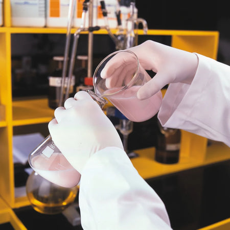 Close-up of a scientist wearing Proval Securitex Premium Latex Examination Gloves while pouring pink liquid between glass beakers in a laboratory environment, demonstrating precision handling and protection.